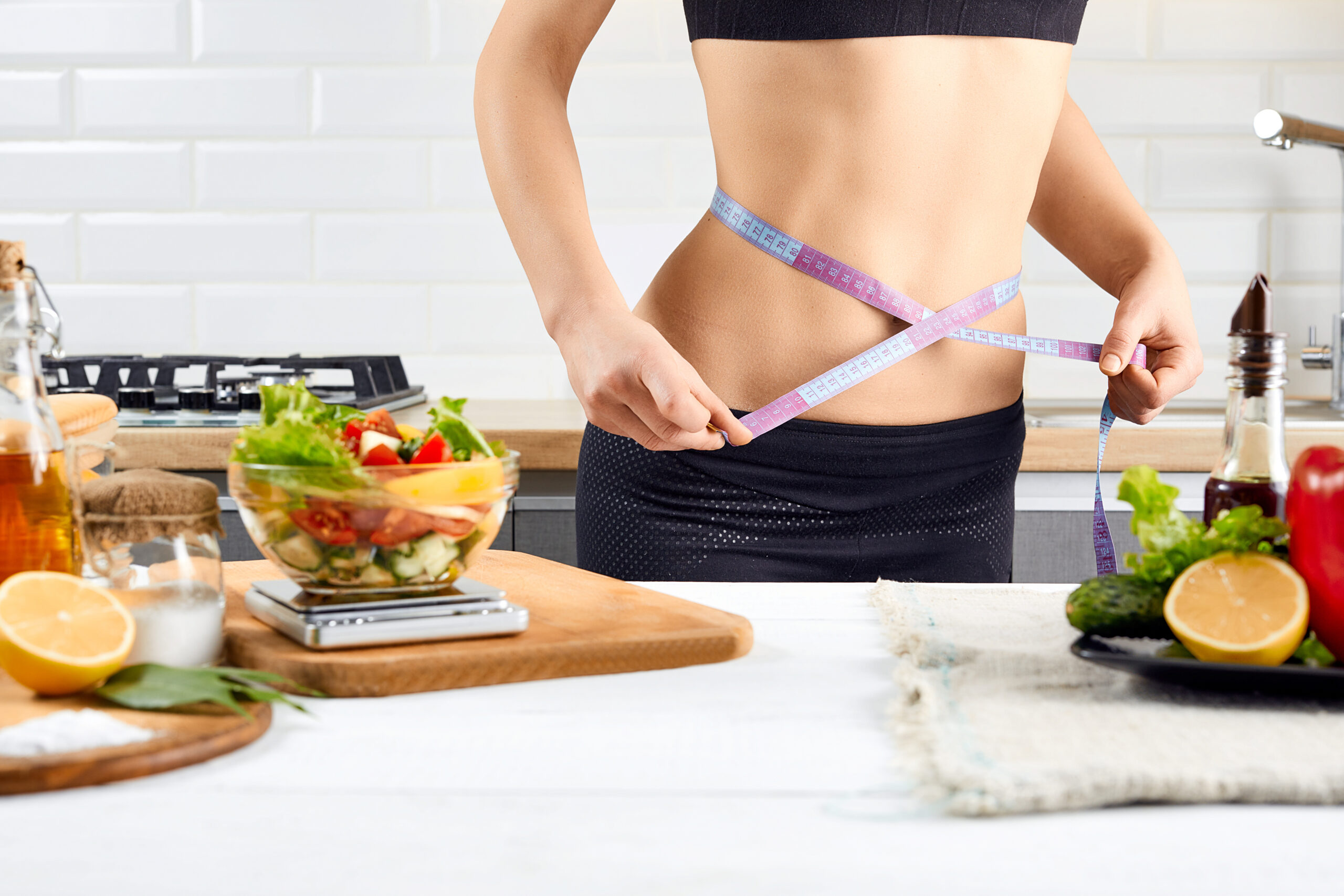 Diet, healthy eating, food and weigh loss concept. Young woman measuring waist near tomatoes, peppers and salad on the kitchen interior.